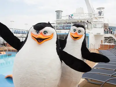 Zwei lachende Pinguin-Maskottchen auf dem Pooldeck der Quantum of the Seas. Ein Swimmingpool, Liegestühle und ihre Aufbauten sind sichtbar.