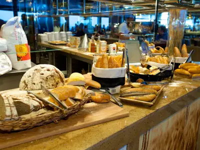 Ein üppiges Buffet mit einer großen Auswahl an frischem Brot und Brötchen. Verschiedene Laibe, darunter ein runder Roggenlaib, sowie Baguettes und Brötchen mit Mohn sind zu sehen. Im Hintergrund sind Mehlsäcke und ein geschäftiger Restaurantbereich erkennbar.
