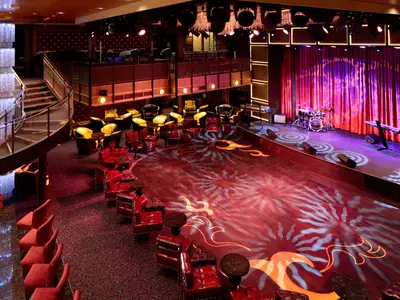 Lounge der Quantum of the Seas. Eine Bühne mit Instrumenten, Tanzfläche, rote und goldene Sitzbereiche, Wendeltreppe und Kronleuchter.
