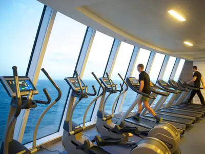 Ein Fitnessstudio an Bord der Quantum of the Seas, ausgestattet mit Ellipsentrainern und Laufbändern. Zwei Männer trainieren mit Blick auf das offene Meer durch Panoramafenster.