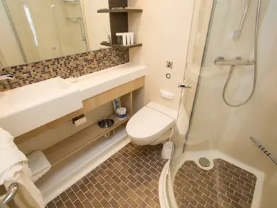 Ein kompaktes Badezimmer an Bord der Quantum of the Seas. Es zeigt einen Waschtisch mit Spiegel, eine Toilette und eine Duschkabine mit Brause und modernem Fliesenboden.