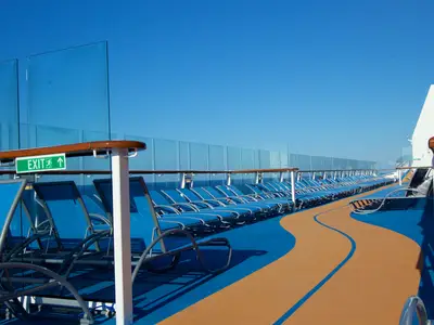 Sonniges Oberdeck der Quantum of the Seas mit Reihen blauer Liegestühle und einer orange-blauen Laufbahn. Ein "EXIT"-Schild ist sichtbar. Der Himmel ist klarblau.