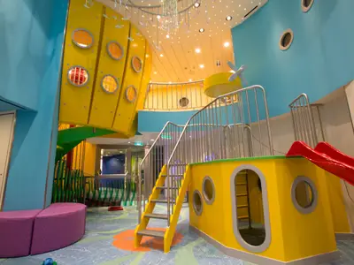 Ein farbenfroher Indoorspielplatz an Bord der Quantum of the Seas. Zu sehen sind gelbe Klettergerüste, eine rote Rutsche, eine grüne Wendeltreppe und eine lila Sitzbank.
