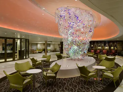 Zentrale Lounge der Quantum of the Seas mit einer großen, schillernden Mosaik-Skulptur. Grüne Sessel, Tische auf Teppich. Rechts eine Bar, links Geschäfte. Orangefarbene Decke.