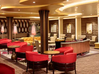 Elegante Lounge auf der Quantum of the Seas mit roten Sesseln, gelben Sofas, beige Stühlen und Holzpaneelen. Warmes Licht betont die geschwungene Decke und Wandkunst.