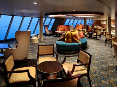 Innenansicht der stilvollen Observation Lounge der Quantum of the Seas. Große Fensterfronten bieten Meerblick, ergänzt durch diverse, farbenfrohe Sitzbereiche und einen gemusterten Teppichboden.