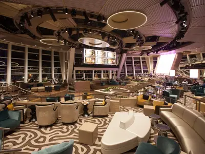 Die geräumige, mehrstöckige Hauptlounge der Quantum of the Seas zeigt eine Vielzahl von Sitzgelegenheiten, große Fenster und eine markante Deckenbeleuchtung. Im Hintergrund sind mehrere Bildschirme sichtbar, während Gäste die Atmosphäre genießen.