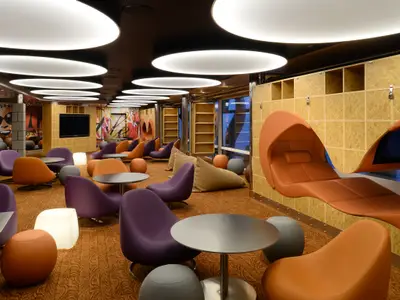 Blick in eine farbenfrohe Lounge auf der Quantum of the Seas, ausgestattet mit modernen lila und orangefarbenen Sesseln, kleinen Tischen und einer futuristischen Liege mit Bildschirm.
