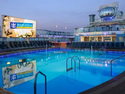 Das Pooldeck der Quantum of the Seas in der Dämmerung mit beleuchtetem Pool, Liegestühlen, Großbildschirm und Sky Bar.