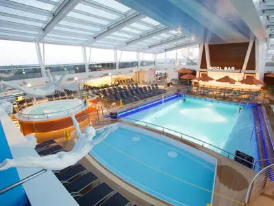 Innenpoolbereich der Quantum of the Seas mit Hauptpool, Lap-Pool und Whirlpool. Liegestühle, eine Poolbar mit Schirmen und weiße Skulpturen sind unter dem Glasdach zu sehen.