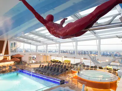 Der überdachte Poolbereich der Quantum of the Seas mit Pool, Whirlpool, Liegen, und mehreren hängenden Skulpturen unter einem Glasdach.