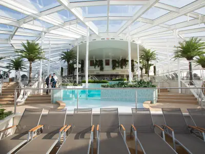Innenansicht des Poolbereichs auf der Quantum of the Seas: Ein Schwimmbecken mit Glaswand, viele Liegen, Palmen und ein helles Glasdach. Personen auf Treppen im Hintergrund.