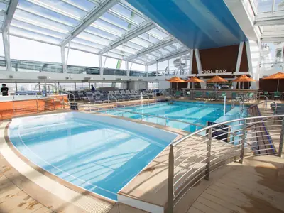 Das Bild zeigt den überdachten Poolbereich der Quantum of the Seas mit zwei Pools, einer Poolbar mit orangenen Schirmen und vielen Liegestühlen unter dem Glasdach.