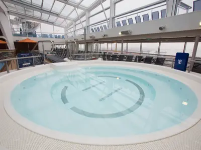 Runder Whirlpool im Solarium an Bord der Quantum of the Seas. Im Hintergrund Liegestühle, Glasdach und Panoramafenster mit Hafenblick.