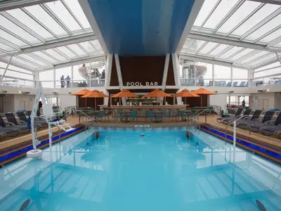 Der überdachte Poolbereich der Quantum of the Seas mit Glasdach, Bar, Liegestühlen und Pool. Ein Poollift und Passagiere auf der oberen Ebene sind sichtbar.