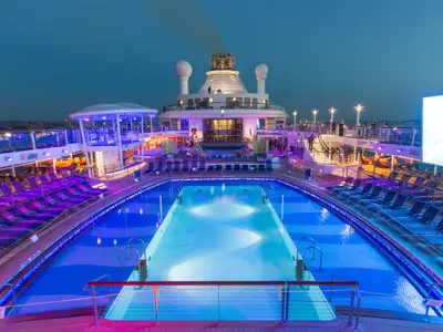 Die Quantum of the Seas bei Nacht: Der beleuchtete Hauptpoolbereich mit blauen und violetten Lichtern und vielen Liegestühlen. Schornstein und Radarkuppeln im Hintergrund.