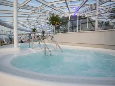 Innenpoolbereich der Quantum of the Seas. Ein sprudelndes Becken mit Edelstahlgeländern unter einem Glasdach. Weiße Flächen, Palmen und Tageslicht prägen das Bild.