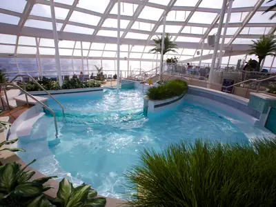 Ein lichtdurchfluteter Indoor-Poolbereich mit blauer Wasseroberfläche, umgeben von grünen Pflanzen und Palmen. Große Glasfenster bieten einen Panoramablick auf das offene Meer. Mehrere Personen entspannen im Hintergrund.