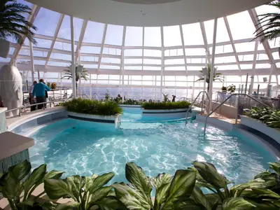 Der helle Solarium-Poolbereich der Quantum of the Seas mit mehreren Becken, umgeben von Pflanzen und Palmen. Glasdach, Meerblick, Personen auf den Decks.