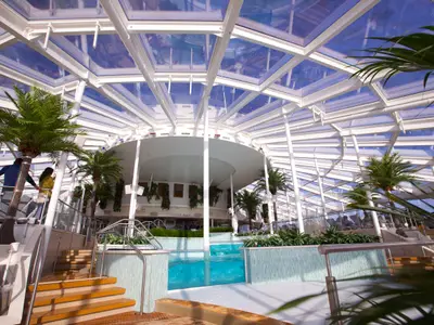 Innenbereich der Quantum of the Seas mit gläsernem Dach. Zu sehen sind Palmen, ein Stufenpool und eine Bar. Wenige Passagiere entspannen in der hellen, modernen Umgebung.
