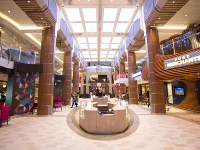 Weitwinkelaufnahme des belebten, mehrstöckigen Innenbereichs der Quantum of the Seas. Man sieht Geschäfte, Restaurants und Sitzgruppen unter einem hellen Dach mit Oberlichtern. Holzverkleidete Säulen strukturieren den Raum.
