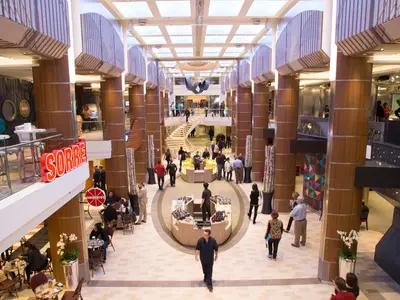 Das belebte Atrium der Quantum of the Seas zeigt Passagiere, Cafés und Geschäfte. Eine Wendeltreppe und zentrale Kioske prägen ihren mehrstöckigen Bereich.