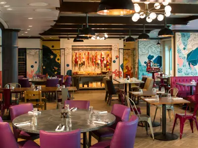 Innenansicht eines modernen Restaurants mit Holztischen, violetten Stühlen und Bänken. Bunte Wandmalereien und ein Feinkostbereich mit hängenden Würsten und Tomaten.