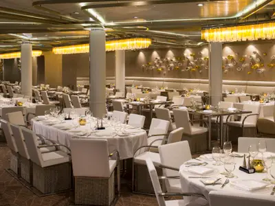 Elegantes Restaurant auf der Quantum of the Seas mit vielen weiß gedeckten Tischen, Stühlen und Sitzbänken. Moderne Wanddekorationen und helle Kronleuchter prägen das Ambiente.