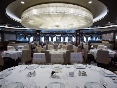 Ein weitläufiger, eleganter Speisesaal der Quantum of the Seas. Viele runde Tische sind mit weißen Tischdecken, Gläsern und Besteck festlich gedeckt. Ein großer, runder, beleuchteter Kronleuchter dominiert die Decke des Raumes.