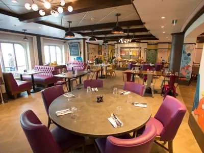 Innenansicht eines farbenfrohen Restaurants der Quantum of the Seas mit lila Stühlen, Holzboden, großen Fenstern, sowie bunten Wandgemälden und einem Buffetbereich im Hintergrund.
