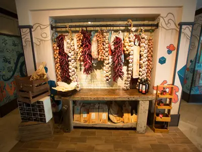 Rustikal eingerichtetes Restaurant mit einer langen Theke aus dunklem Holz. Darüber hängen Zöpfe aus roten Chilischoten, Knoblauch und Zwiebeln. Links stapeln sich Holzkisten, unter der Theke sind Holzschneidebretter. Die Wände sind mit abstrakten Mustern in Blau und Orange verziert. Keine Personen zu sehen.
