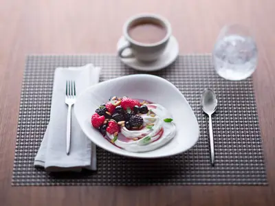 Eine nahaufnahme eines reichhaltigen Frühstücks mit Joghurt, frischen Himbeeren, Brombeeren, Heidelbeeren und Pinienkernen in einer weißen Schale. Daneben liegen eine Gabel und ein Löffel, im Hintergrund sind eine Kaffeetasse und ein Wasserglas unscharf zu sehen, alles auf einem Holztisch mit geflochtenem Tischset.