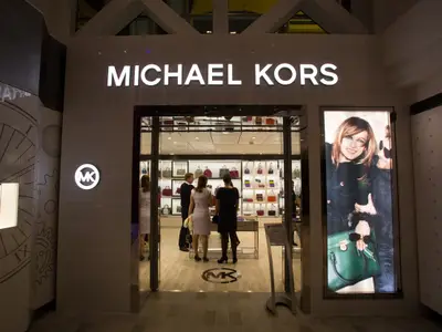 Eingang eines Michael Kors Geschäfts auf der Quantum of the Seas. Beleuchtetes Logo, Taschenauslage und Werbeplakat mit Frau und grüner Tasche.