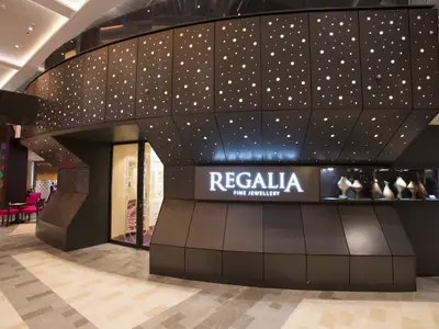 Innenansicht der Quantum of the Seas. Das Juweliergeschäft 'Regalia Fine Jewellery' mit beleuchtetem Logo, dunkler, punktierter Fassade. Schaufenster mit Skulpturen und Sitzbereich sind erkennbar.