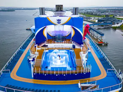 Luftaufnahme der Quantum of the Seas im Hafen, mit einer Person auf dem FlowRider-Surfsimulator am Heck. Rechts ein großer pinker Bär. Im Hintergrund ist eine Stadt zu sehen.