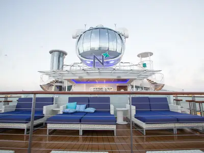 Oberdeck der Quantum of the Seas mit gläserner North Star Kapsel, N-Logo. Lounge-Möbel mit blauen Polstern auf Holzdeck, Reling im Vordergrund. Bewölkter Himmel.