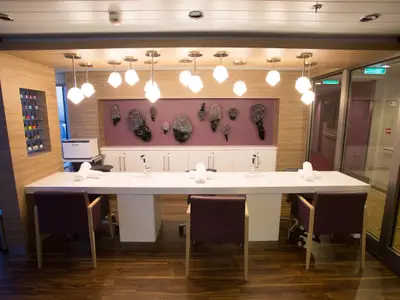 Modernes Nagelstudio an Bord der Quantum of the Seas. Weiße Theke mit Waschbecken, Stühlen, Handtüchern. Lila Wand mit Kunstobjekten und Hängelampen.