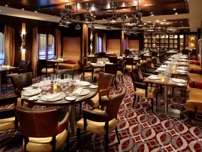 Restaurant auf der Quantum of the Seas mit vielen gedeckten Tischen. Man sieht braune Ledersessel, lange Sitzbänke, moderne Leuchten und einen roten Teppich.