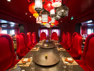 Innenansicht eines asiatisch inspirierten Spezialitätenrestaurants. Lange Tische mit roten Samtstühlen und bunten Deckenlampen in Form von Laternen. Am Ende des Raumes sind Fenster mit roten Vorhängen zu sehen. Die Wände sind ebenfalls in einem dunklen Rotton gehalten.