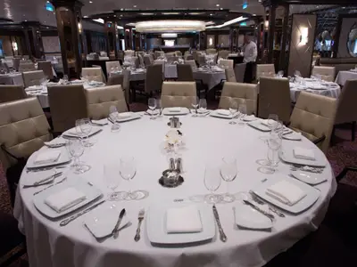 Ein großer, eleganter Speisesaal an Bord der Quantum of the Seas mit vielen runden, weiß gedeckten Tischen. Jeder Tisch ist mit Geschirr, Besteck und Gläsern für mehrere Personen ausgestattet. Im Hintergrund sind weitere Tische und ein Mitarbeiter zu sehen.