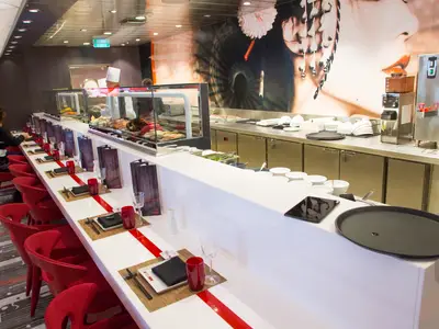 Sushi-Restaurant der Quantum of the Seas. Lange Theke mit roten Hockern, gedeckte Plätze. Koch und Speisenvitrine im Hintergrund. Große Kunstwand.