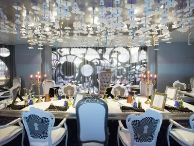 Innenansicht eines opulent dekorierten Restaurants mit langer, weißer Tafel, vielen Stühlen mit Schachbrettmuster und einer Decke voller Schlüssel und pilzförmigen Lichtern.