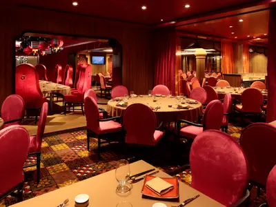 Ein festlich rotes Restaurant an Bord der Quantum of the Seas. Man sieht zahlreiche Tische mit roten Samtstühlen, einen bunten Teppich und Vorhänge. Die Tische sind gedeckt.