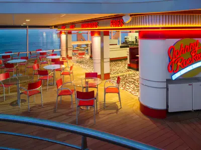 Der beleuchtete Außenbereich des Johnny Rockets Restaurants an Bord der Quantum of the Seas bei Dämmerung, mit roten Stühlen, Glastischen und Blick aufs Meer. Das neonfarbene Logo des Restaurants ist prominent zu sehen.