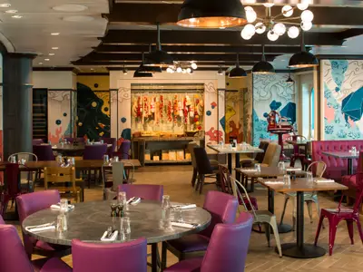 Innenansicht eines farbenfrohen Restaurants an Bord der Quantum of the Seas. Mehrere Tische mit Stühlen, darunter lila gepolsterte Sessel, füllen den Raum. Die Wände sind mit abstrakten, bunten Wandgemälden verziert, und an einer Theke hängen Wurstwaren.