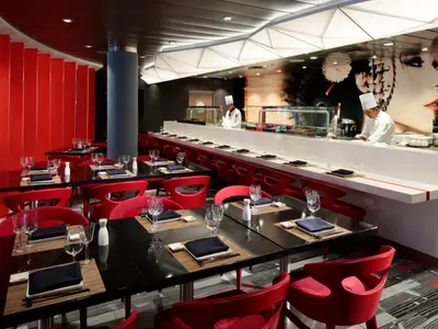 Modernes asiatisches Restaurant auf der Quantum of the Seas. Rote Stühle, schwarze Tische und eine Sushi-Bar mit Köchen. Wandbild, beleuchtete Decke und gedeckte Tische sind sichtbar.