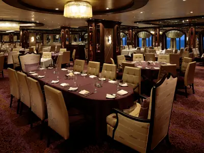 Ein elegantes Restaurant an Bord der Quantum of the Seas. Zahlreiche Tische sind gedeckt, umgeben von gepolsterten Stühlen. Im Hintergrund sind Fenster mit Meerblick sichtbar.
