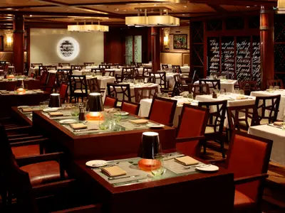 Elegantes Restaurant auf der Quantum of the Seas. Gedeckte Tische, dunkle Holzstühle, gedämpftes Licht und Wanddekorationen.