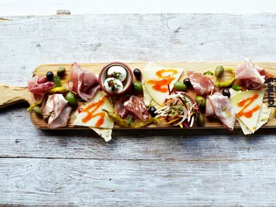 Rustikale Antipasti-Platte mit Wurst, Käse, Oliven und Mozzarella auf einem Holzbrett. Darunter ein weiß gestrichener Holztisch. Logo 'Jamie's Italian'.
