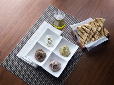 Eine weiße viereckige Schale mit vier verschiedenen Dips und ein Korb mit gegrilltem Fladenbrot und Olivenöl auf einem Holztisch mit geflochtener Unterlage.